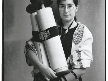 Full body photograph of a person in glasses holding scroll with left arm wrapped in leather strap. 