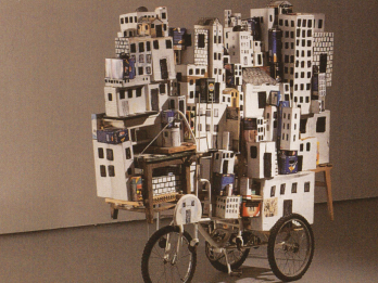 Mixed media work featuring stack of buildings on bicycle. 