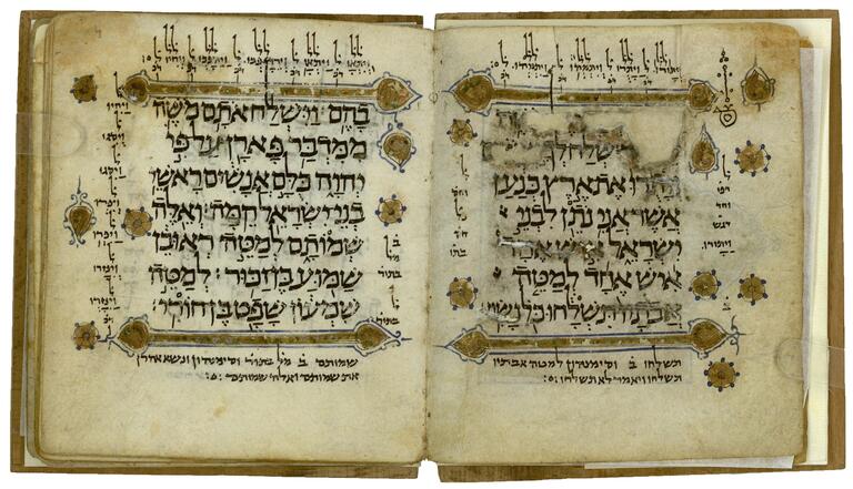 Book open to two facing pages, containing large Hebrew with gold borders above and below, and gold geometric and floral decorations with blue outlines and accents alongside, all surrounded by smaller, vowelless notes and a diagram.