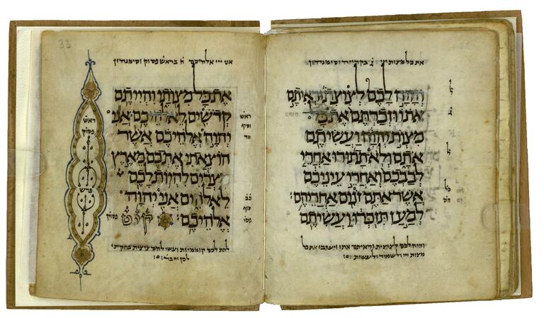 Book open to two facing pages containing large Hebrew writing, with a decorative blue and gold image to the left containing a few small Hebrew words, the final word in outline preceded by a small circular decoration, and smaller, vowelless notes in the margins.