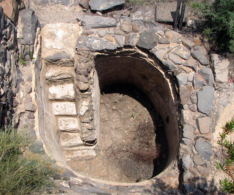 A round, stone-lined pit with built-in stone steps leading down to a dirt floor. The structure is surrounded by rocks and sparse vegetation.