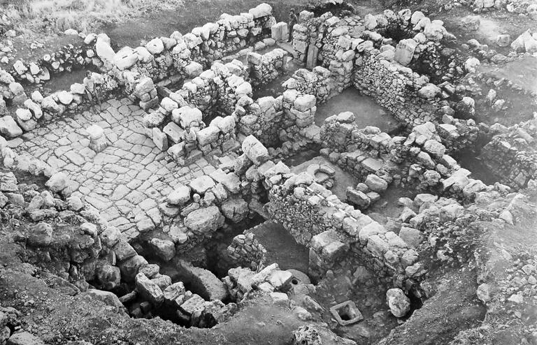 Aerial view of stone ruins, showing multiple rectangular rooms and passageways with uneven stone walls and floors, partially surrounded by earth and vegetation.