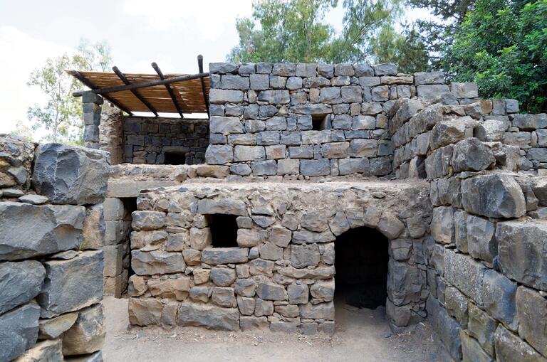 Ancient stone building with small windows and doorways, constructed from large, rough-hewn rocks, surrounded by trees. The structure has multiple levels and is partially covered by a thatch roof.