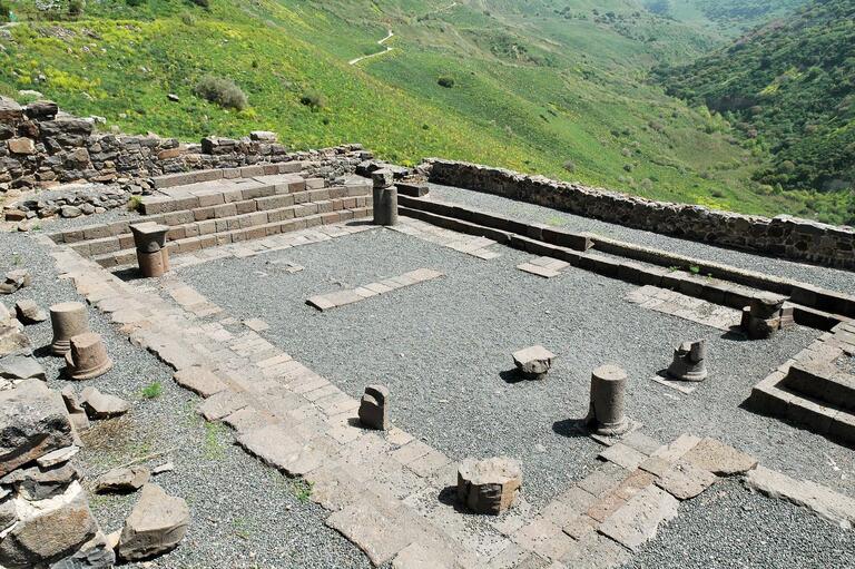 Ruins of an ancient stone structure sit on a gravel surface, surrounded by low stone walls and scattered columns, overlooking a lush, hilly landscape.