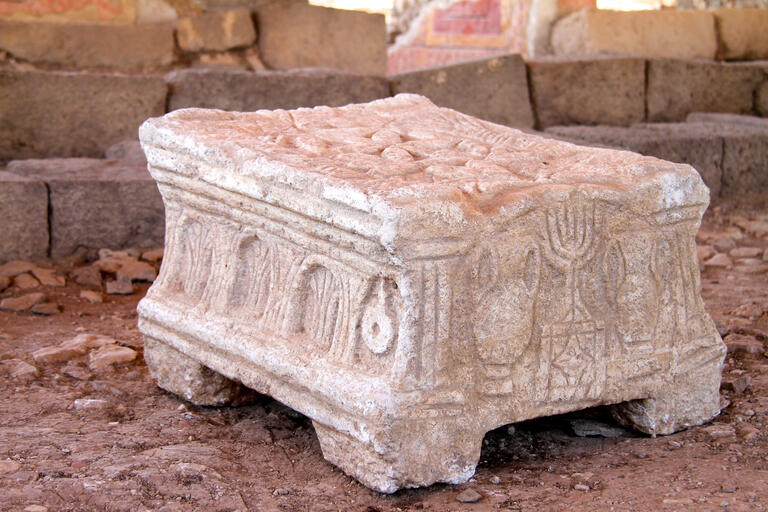 A carved stone object with detailed reliefs, including menorahs and arches, resting on four short legs, sits on a dirt floor among weathered stone blocks and ruins.