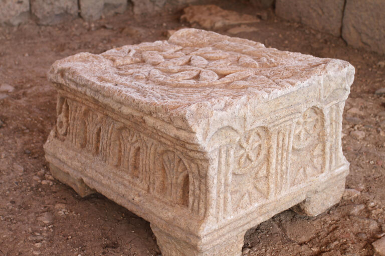 A carved stone object with ornate patterns and symbols, including rosettes or wheels within arches, resting on four short legs, sits on a dirt floor among weathered stone blocks and ruins.