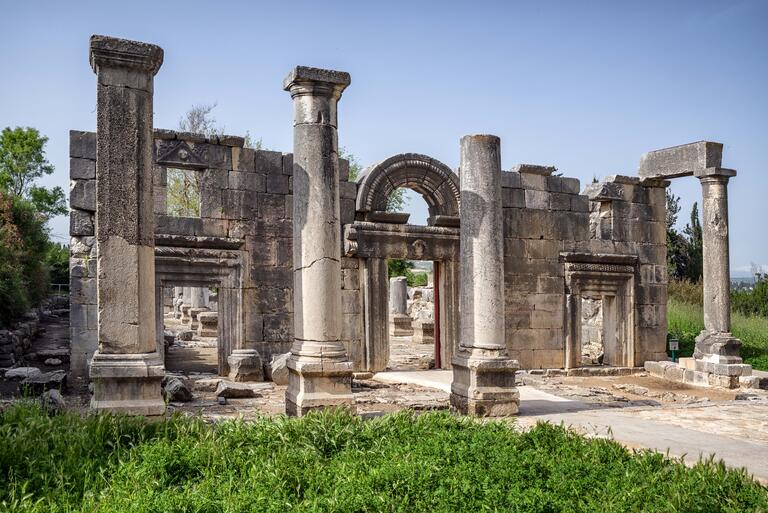 Partially preserved stone building facade with an arched entrance and four columns in front. Other stone ruins are visible through the entryway.