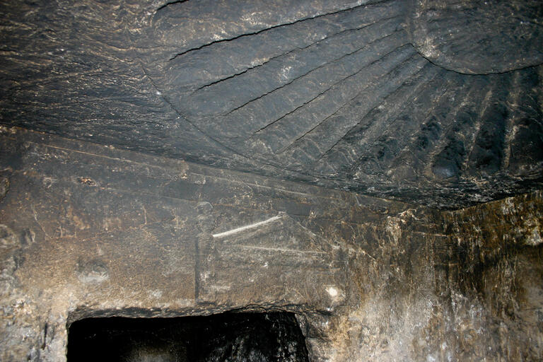 A stone ceiling with a large, fan-shaped relief carved into it, above a rough doorway in an ancient, dark stone chamber. The walls are textured and worn.