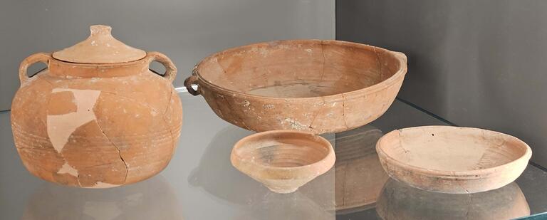 Four ancient, weathered ceramic vessels: a lidded jar with handles, a large bowl, a small bowl, and a shallow plate, displayed on a glass shelf against a plain, neutral background.