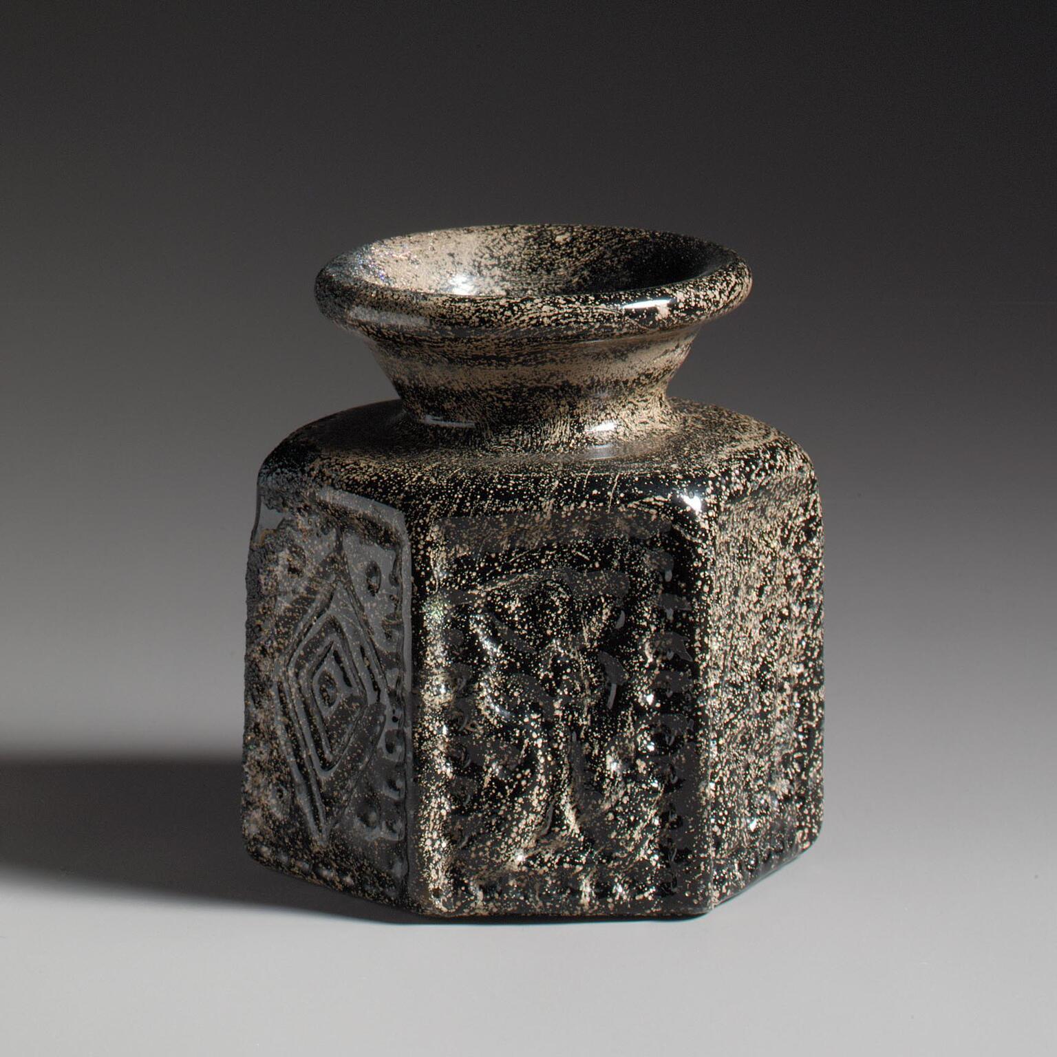 Photo of a dark glass vase with a wide, flared rim and hexagonal base, featuring geometric patterns.