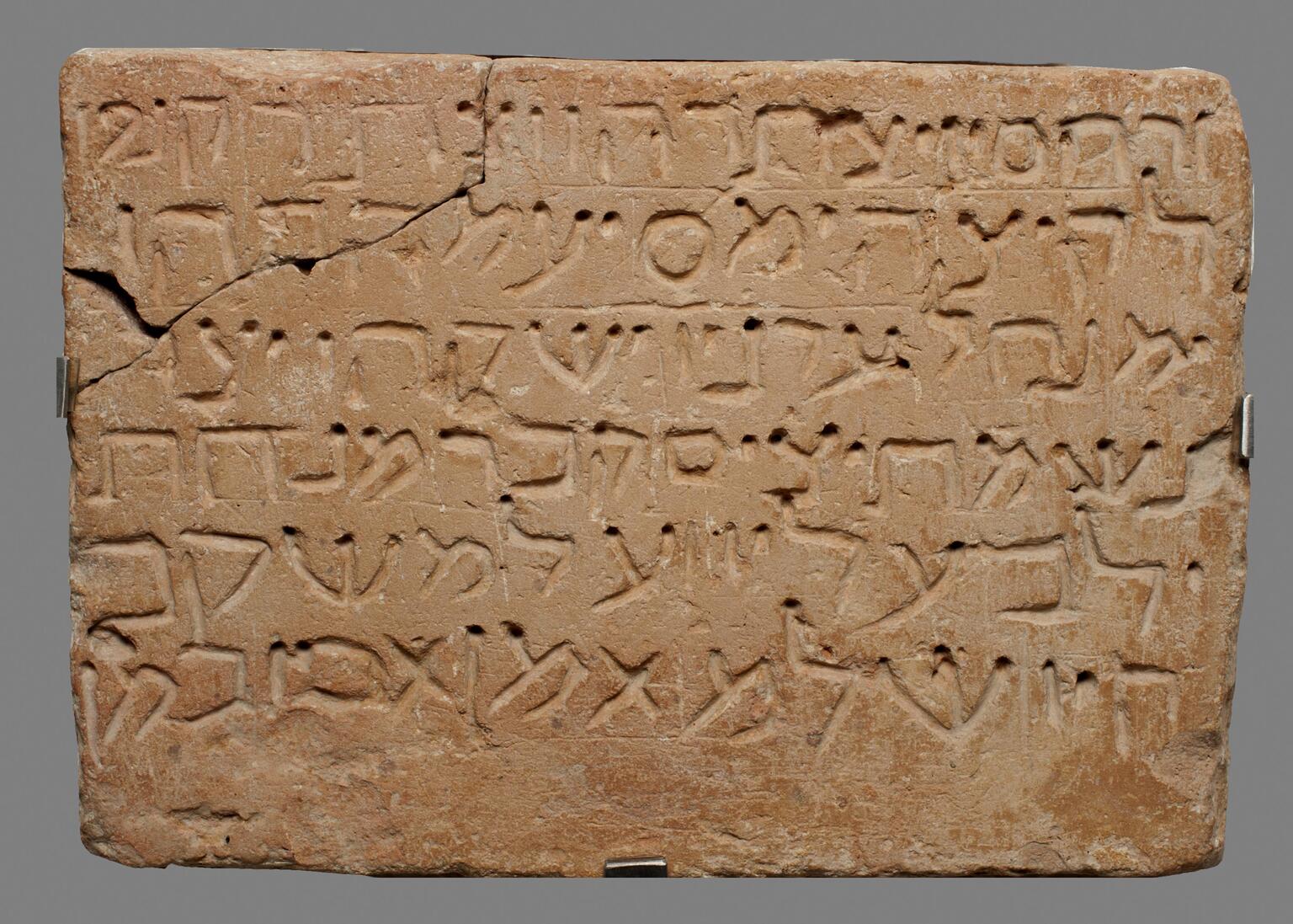 Rectangular brick tablet with six lines of carved Hebrew script and a broken upper-left corner.