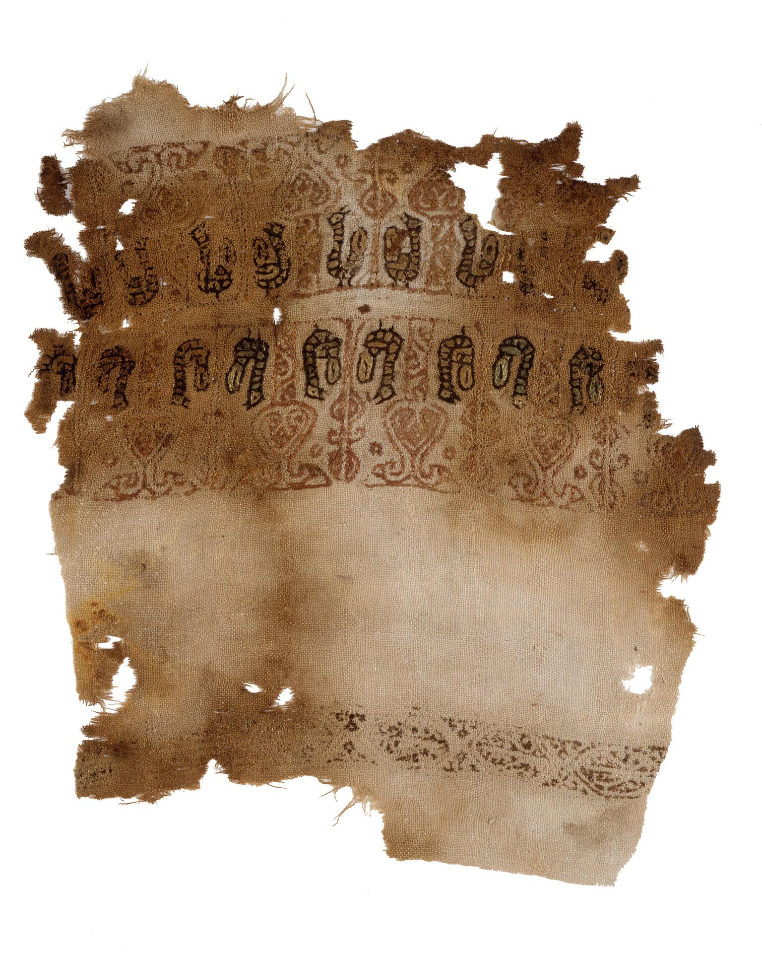 Damaged fragment of fabric with decorative patterns and symbols in dark ink and faded color, mainly red, toward the top and in one additional stripe at the bottom.