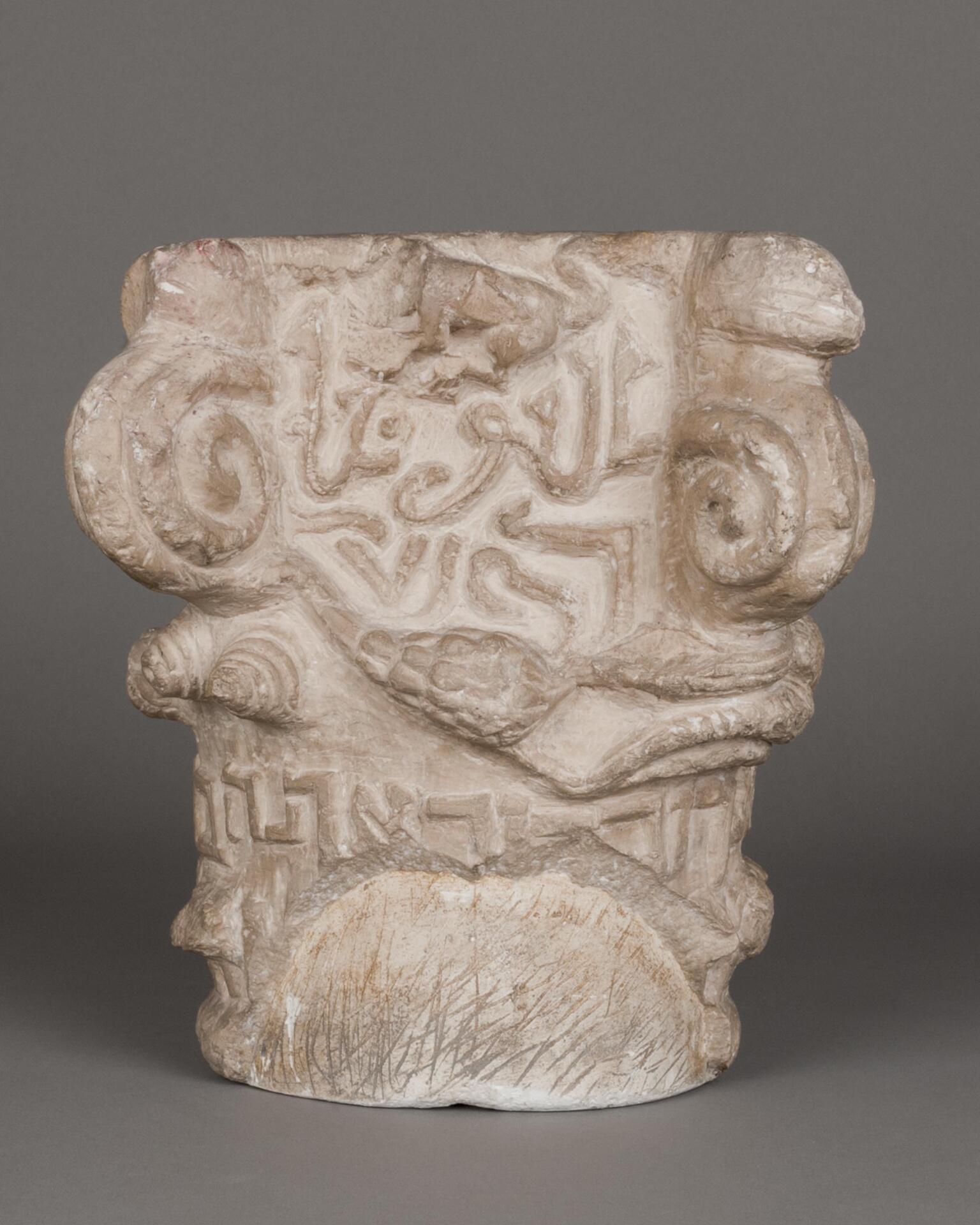 Worn carved stone capital featuring spirals, patterns, flora and fauna, and lines of Arabic and Hebrew lettering.
