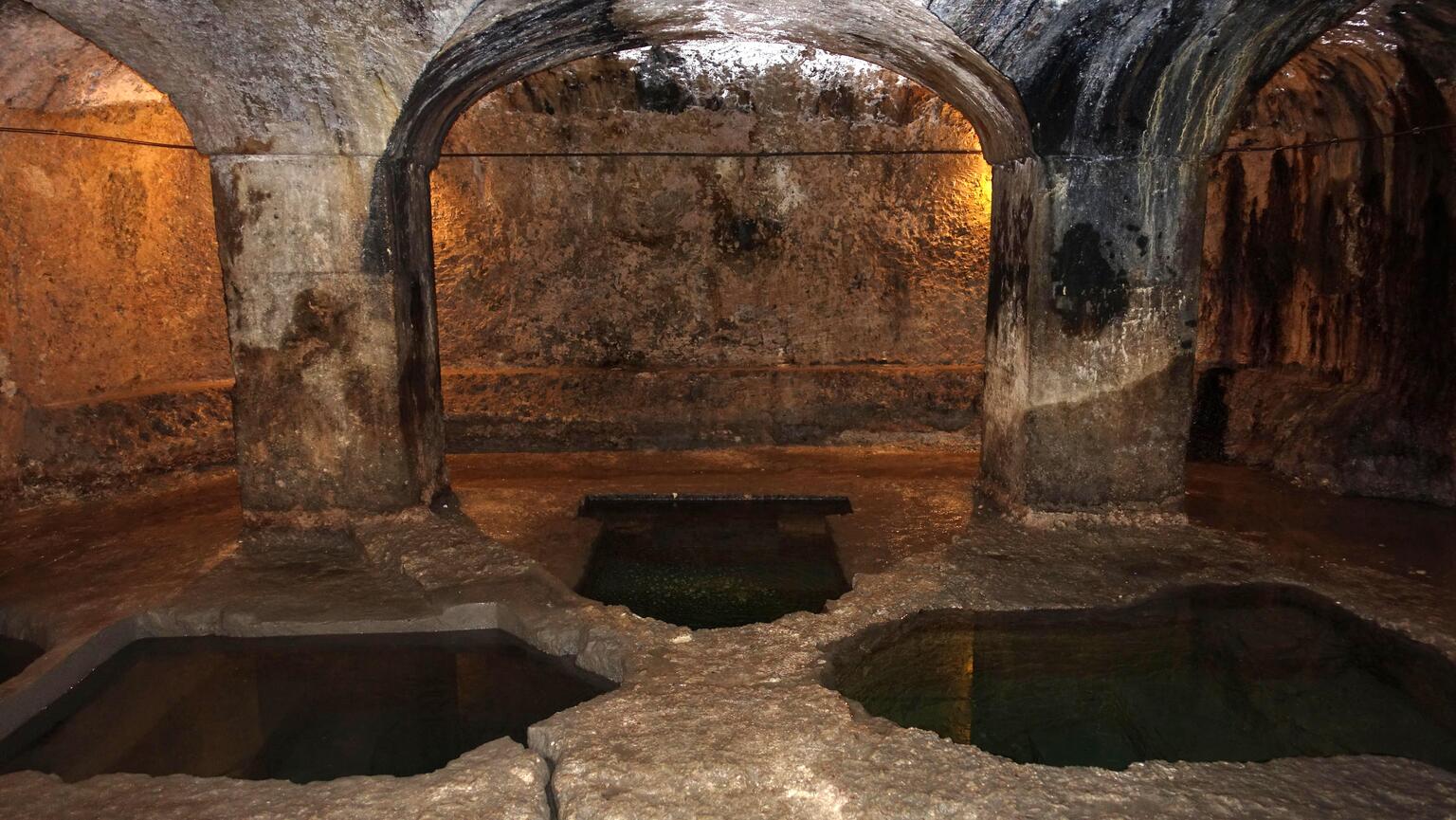 Photo of stone room with arched ceilings, pillars, rough walls, and several pointed pentagonal pools set into the floor.
