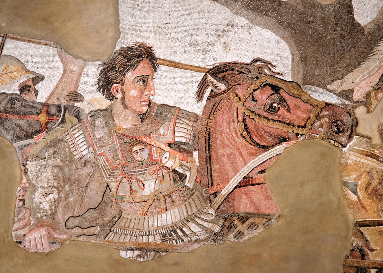 Ancient mosaic depicting a warrior in ornate armor, holding a spear, riding a horse into battle. The warrior has wavy hair and a determined expression and is surrounded by other soldiers. The background is partially damaged.