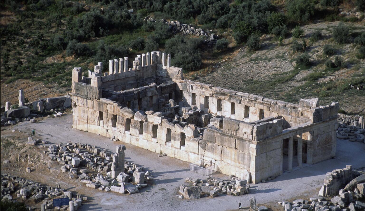 Aerial view of the ruins of an ancient stone building with columns and walls partially standing, surrounded by scattered stones and greenery in the background.