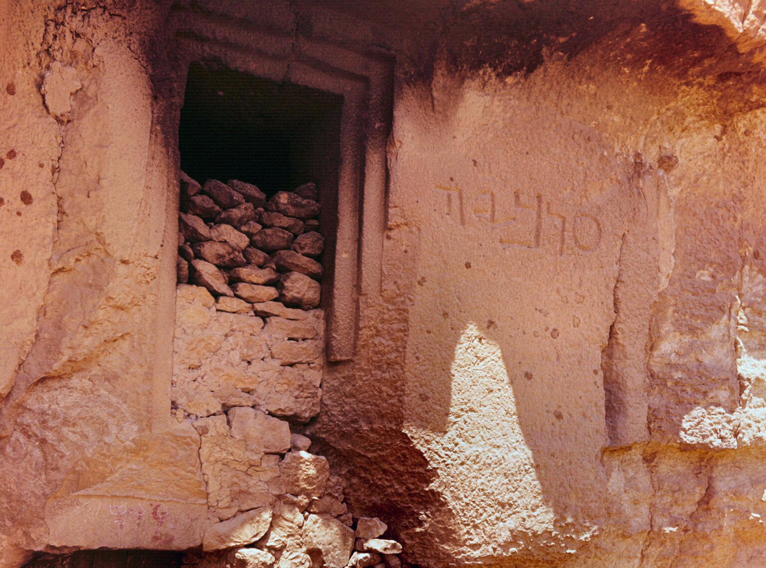 A stone doorway carved into a rock wall, filled with loose stones. There is an inscription to the right of the doorway. The rock surface is rough and bathed in sunlight.