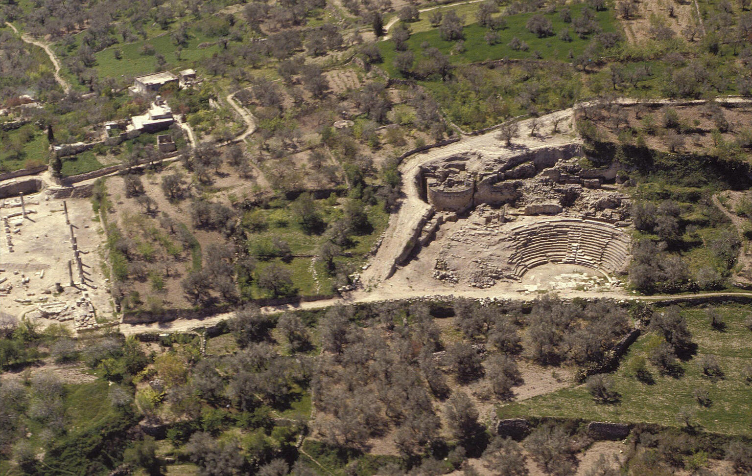 Aerial view of an ancient stone amphitheater surrounded by green fields, scattered trees, and ruins of other structures in a rural landscape.