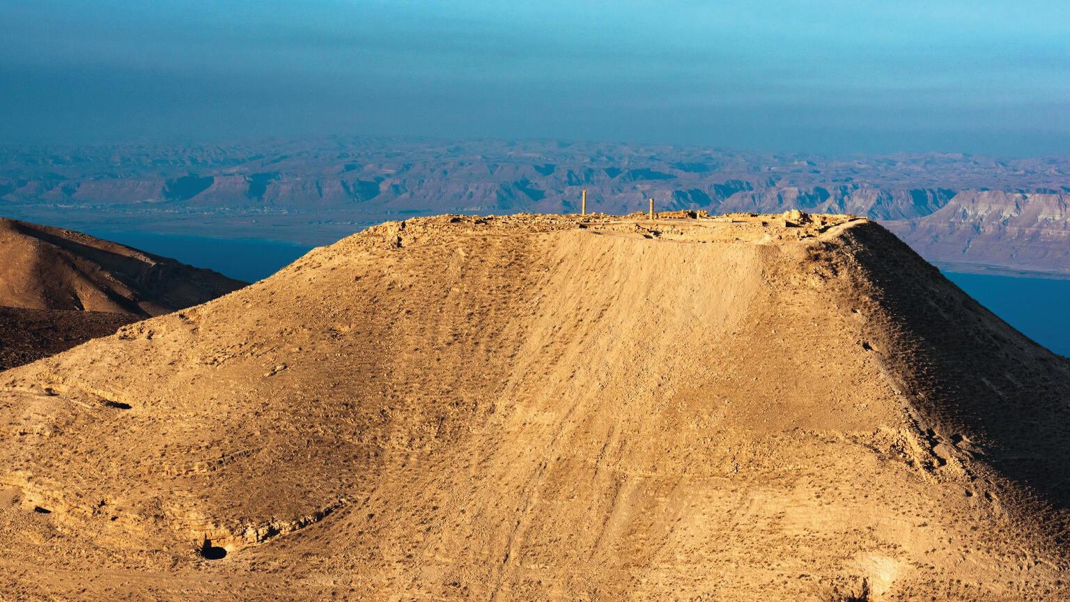 Aerial view of mountain with ruins on top, surrounded by desert landscape, and a sea in the background.