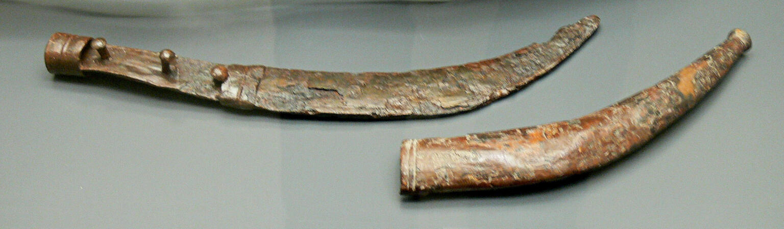 An ancient, curved metal dagger and scabbard displayed side by side on a flat surface. The dagger has a corroded blade and worn surface, and a hilt with three rounded knobs.