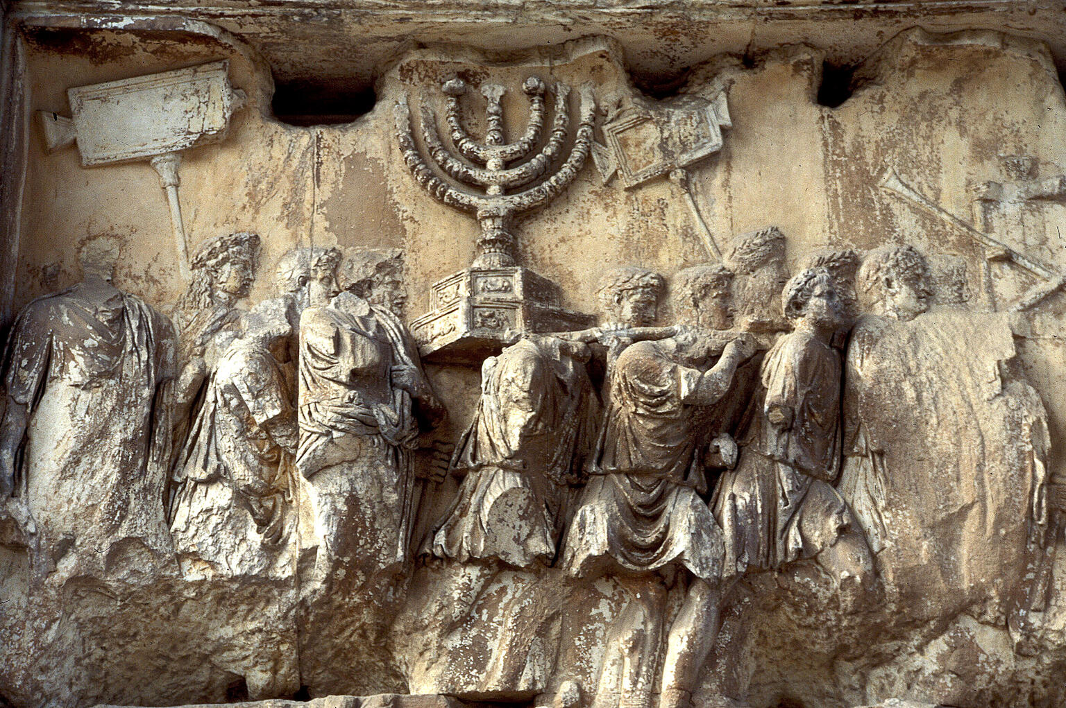 Ancient, weathered stone relief showing people in robes carrying a large menorah and other objects in a procession.