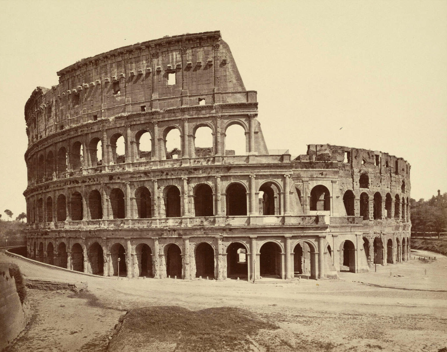 Black-and-white photo of a large amphitheater, showing its ancient stone arches and partial ruin. The large amphitheater stands alone, with some missing sections and an empty foreground.