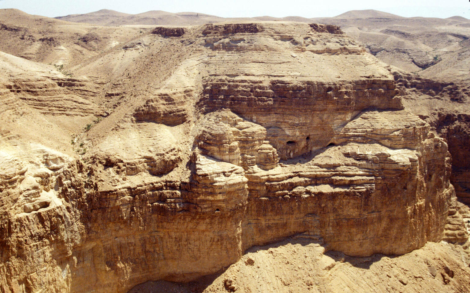 A rugged, sunlit desert canyon with steep, layered rock formations and barren, rolling hills in the background.
