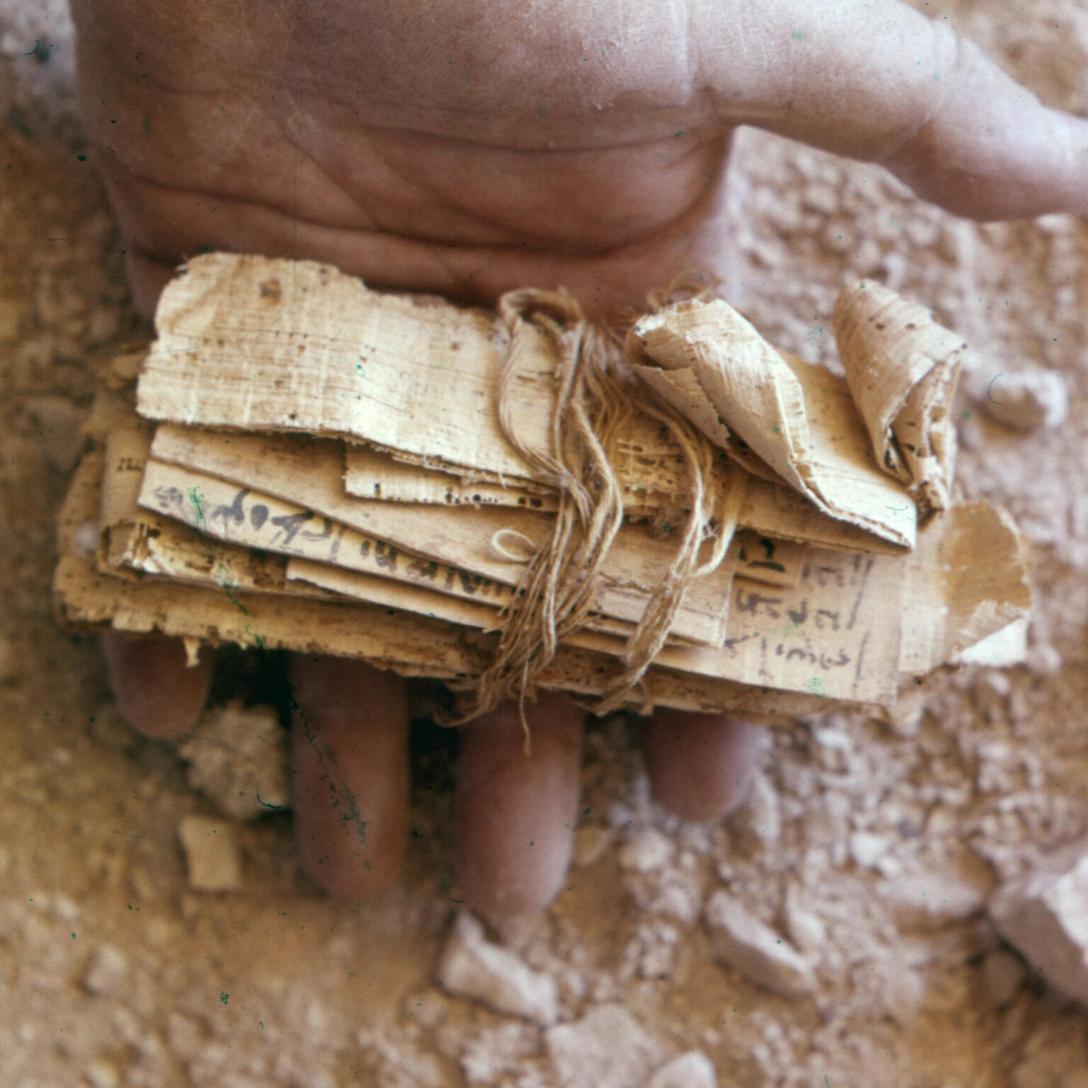 Close-up of a hand holding an ancient, rolled bundle of papyrus tied with string, resting on a dusty surface with small rocks or pieces of debris scattered around.