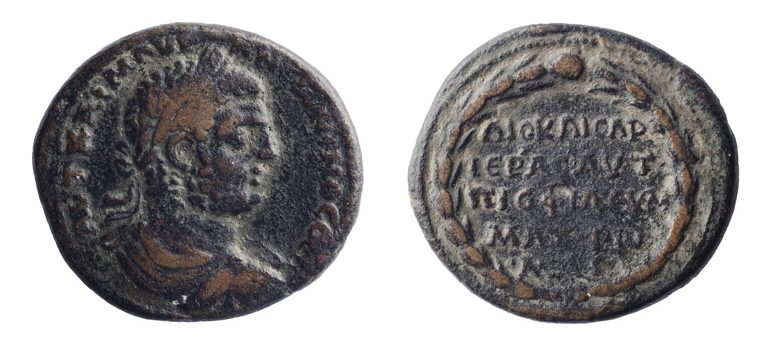 Ancient coin with a profile of an emperor on one side and inscribed Greek text surrounded by a laurel wreath on the other. The coin appears worn and dark in color.