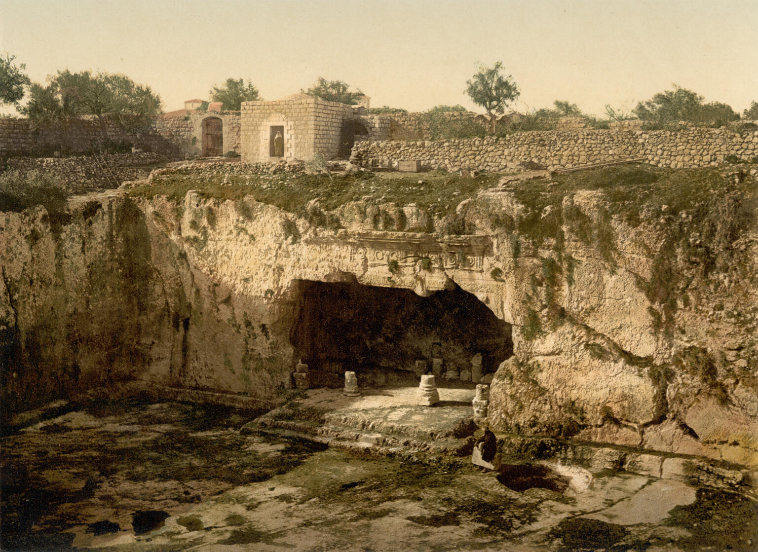 A large ancient rock-cut tomb with pillars and carvings set into a cliff, surrounded by grassy ground and stone walls, with small stone buildings and trees in the background.