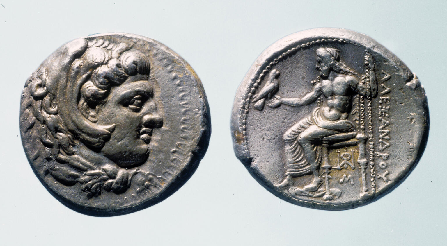 Front and back of an ancient silver coin. One side shows a profile of a man's head with curly hair, while the other side depicts a seated figure holding a bird, with Greek inscriptions beside the figure.