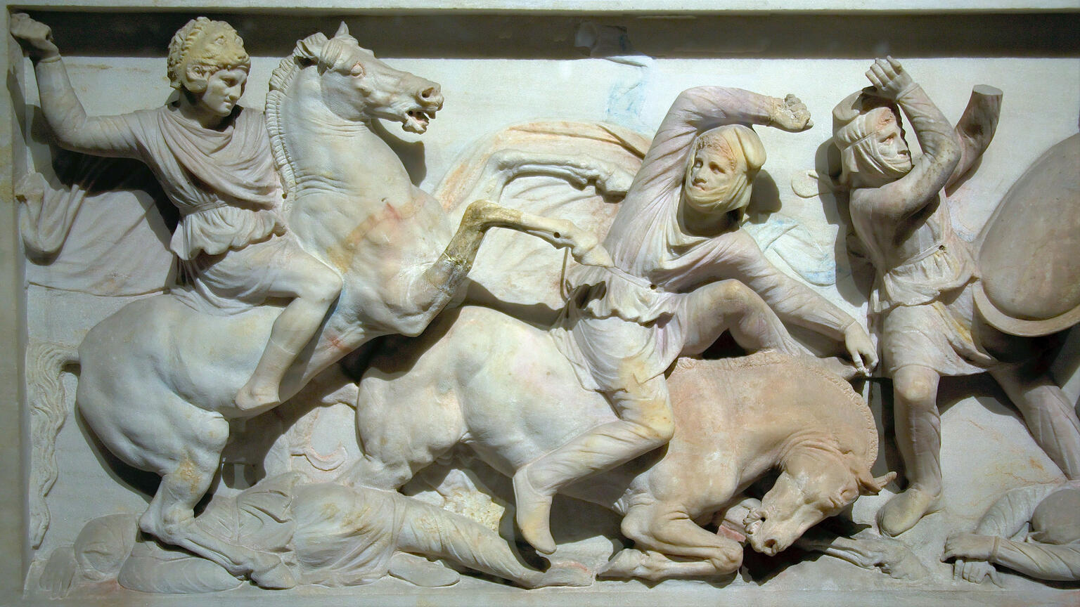 A marble relief sculpture shows three figures in battle: a mounted warrior attacks another figure who is falling from a horse, while a third figure takes a defensive pose. Two additional figures, apparently dead, lie on the ground.