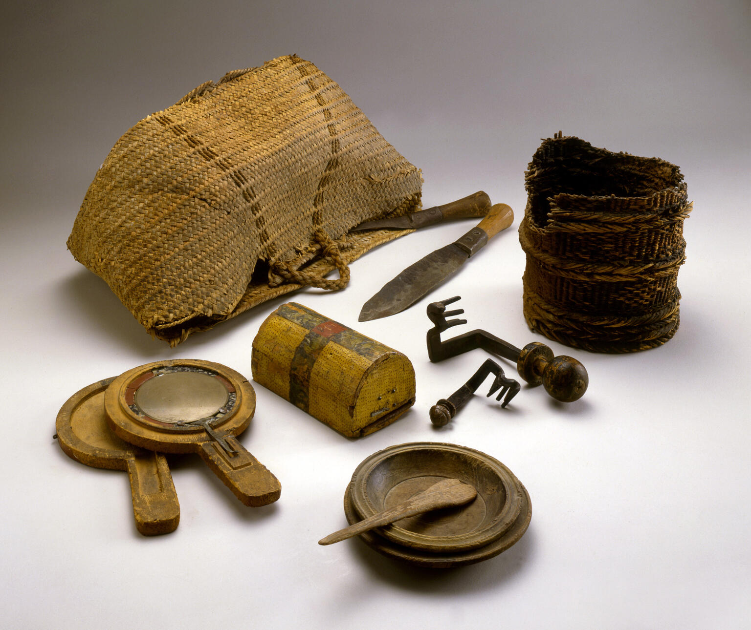 A collection of old household items including woven baskets, a wooden box, knives, keys, handheld mirrors, a wooden bowl with a spoon, and a wooden plate, arranged on a plain background.
