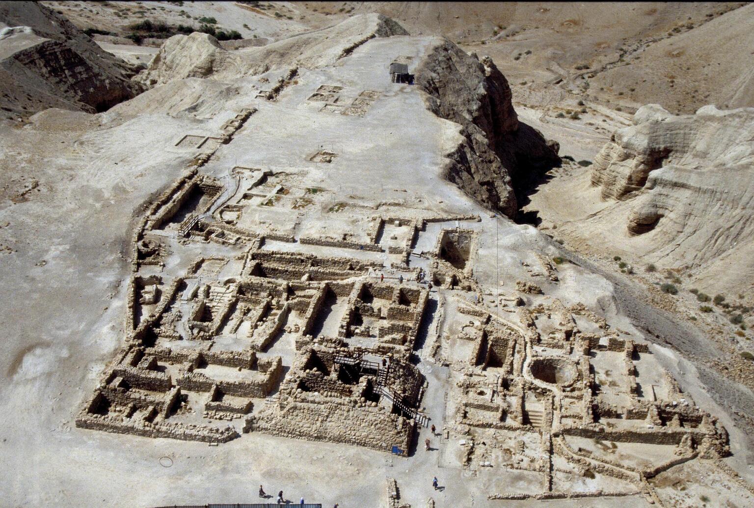 Aerial view of ancient stone ruins in a desert landscape, showing rectangular foundations, walls, and pathways amid rocky terrain and sandy hills. People are visible exploring the site.