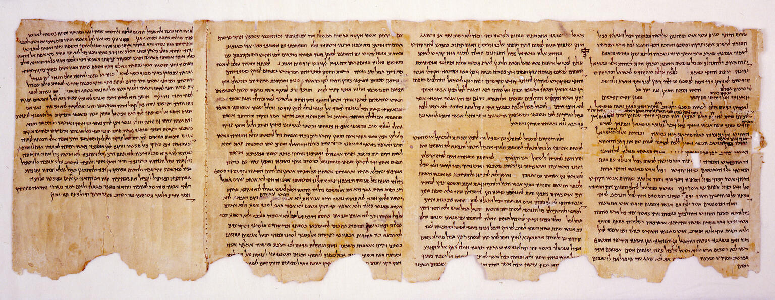 A fragment of an ancient scroll with tightly spaced, dark Hebrew text in horizontal lines on aged, yellowed parchment. The edges are irregular and damaged, with portions missing and discoloration visible.