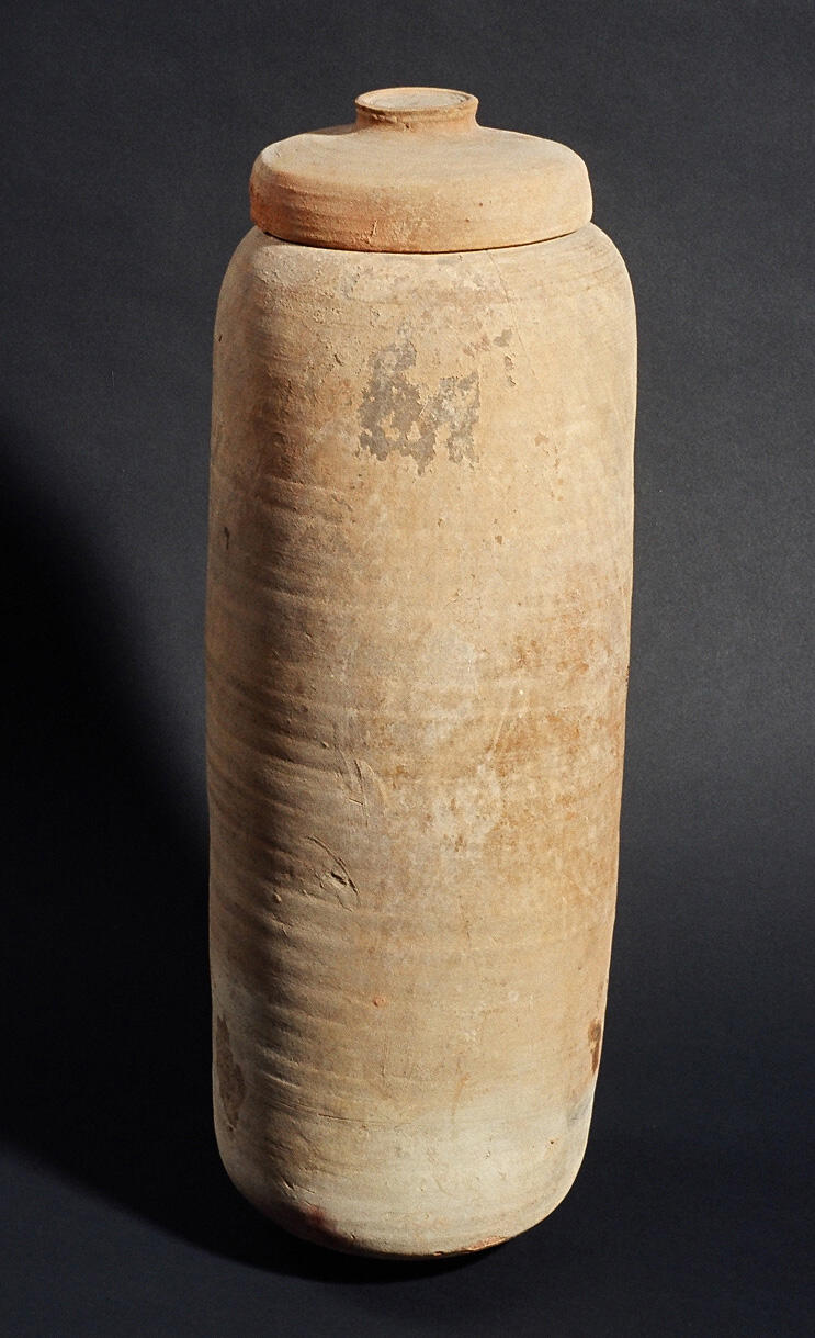 A tall, cylindrical clay jar with a rounded lid, standing upright against a dark background. The surface appears rough and slightly uneven, with some discoloration and markings.