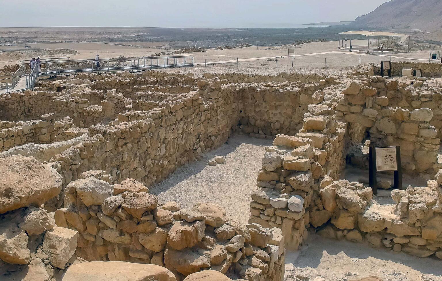 Ancient stone ruins with partially intact walls in a desert landscape. Distant visitors walk on a modern pathway under a hazy sky.