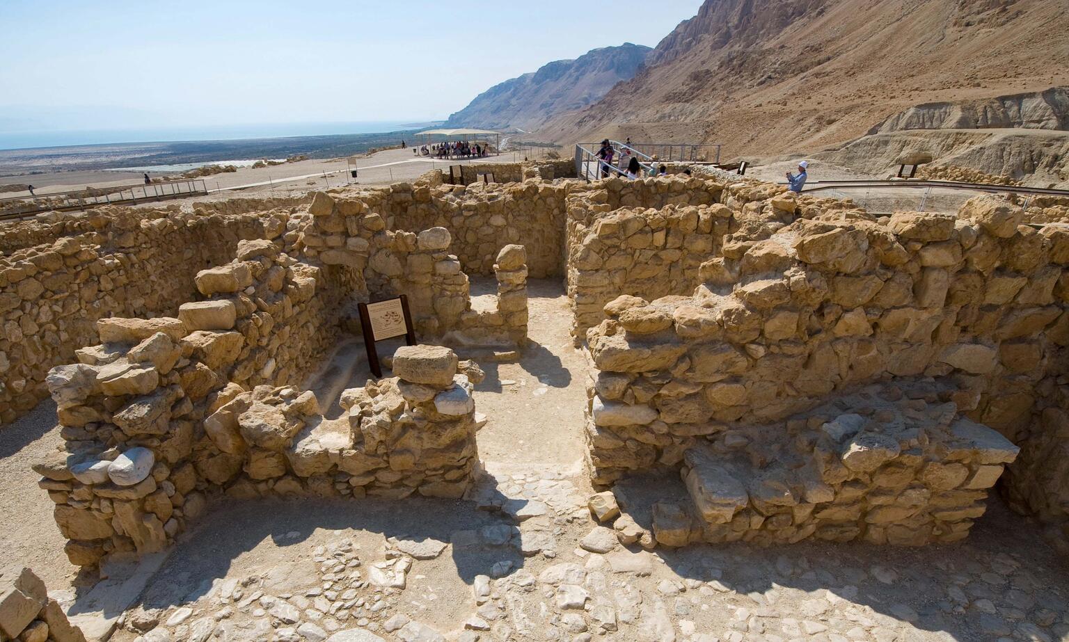 Ancient stone ruins with low walls and doorways under a clear sky, with mountains and the Dead Sea visible in the background. Tourists walk along a pathway nearby.