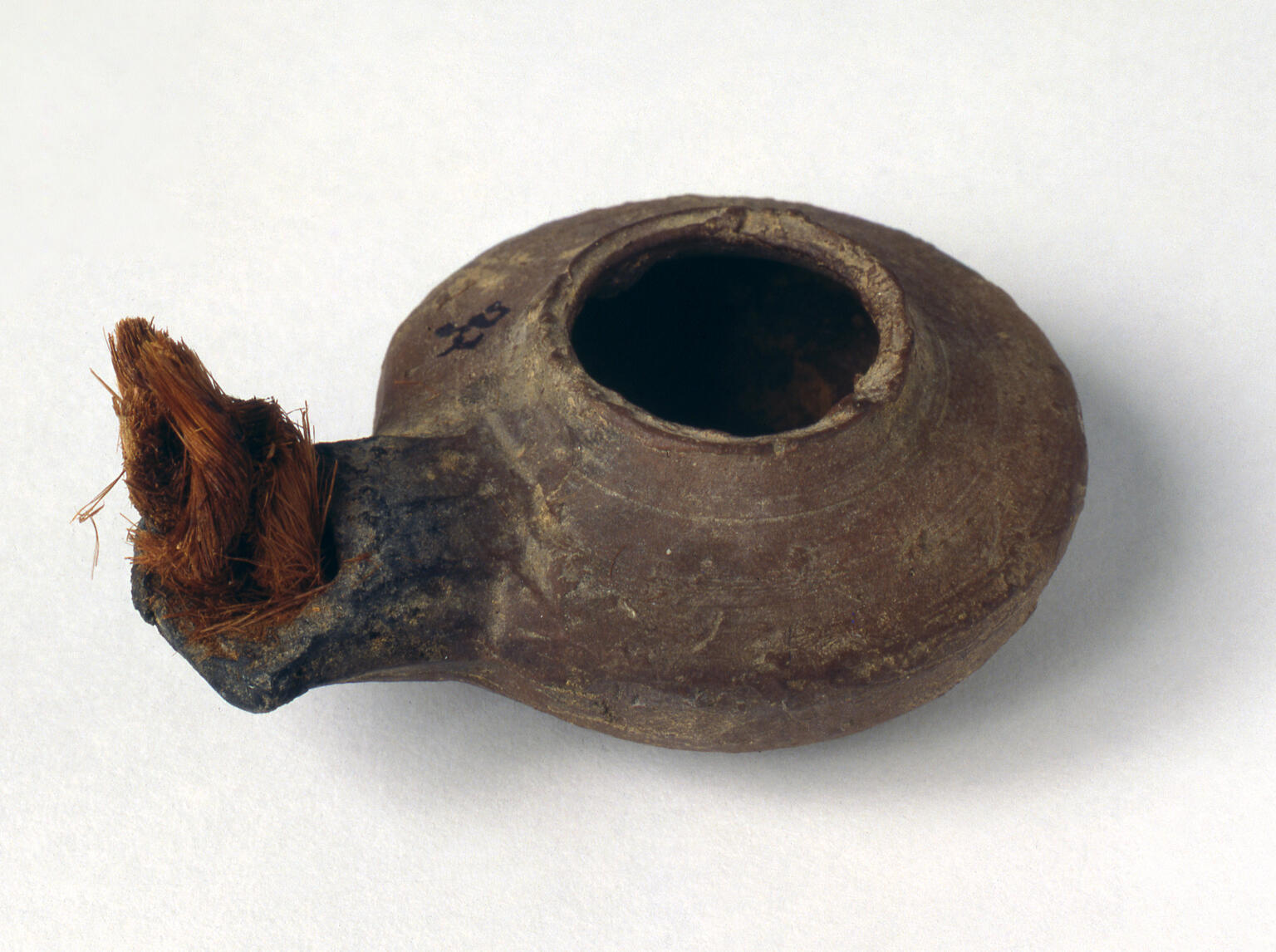 A small, round ancient clay oil lamp with a wide opening, featuring a blackened spout with a fibrous wick inside.