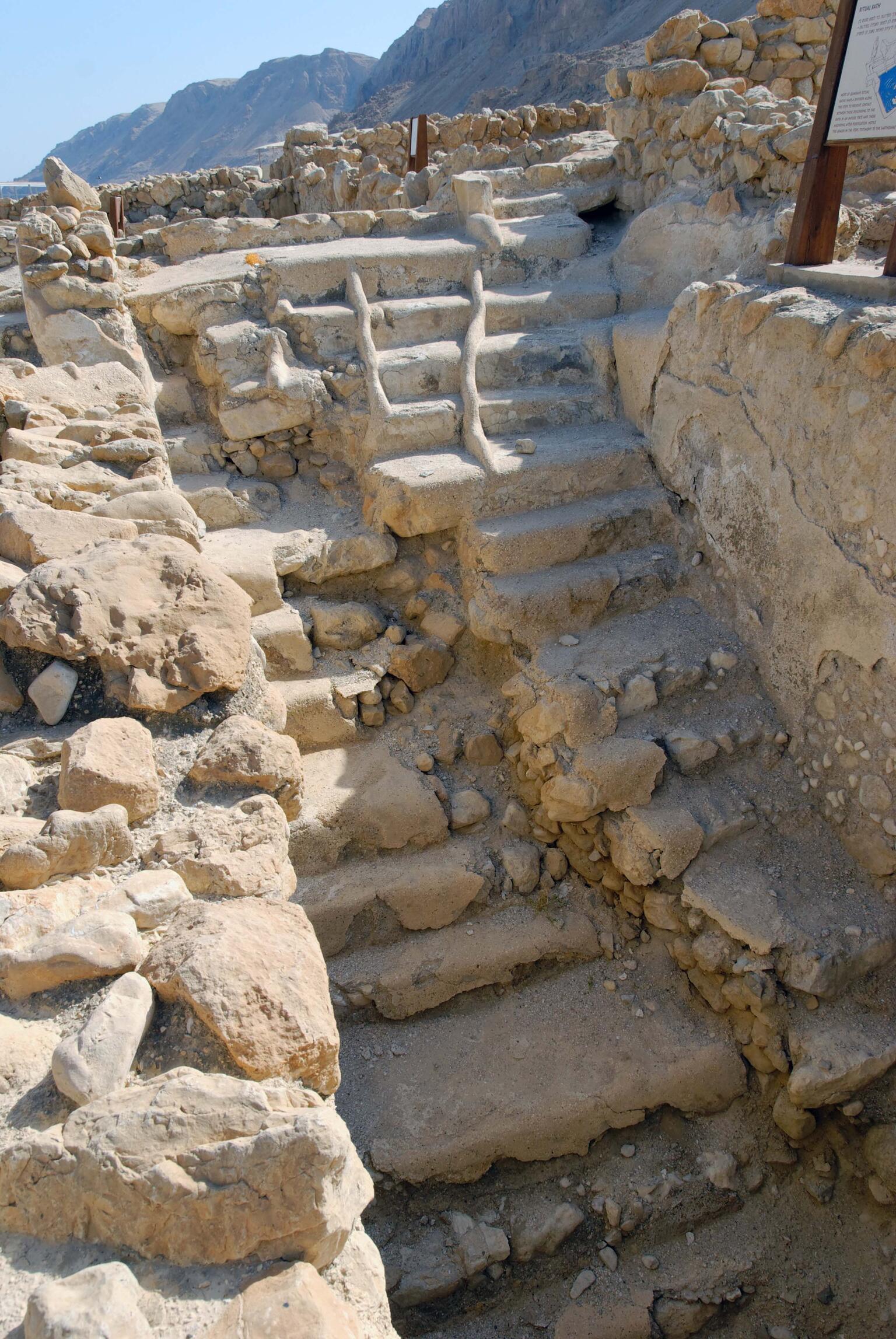 Ancient stone steps built into a rocky structure, surrounded by rough stone walls, with mountains visible in the background under a clear sky.