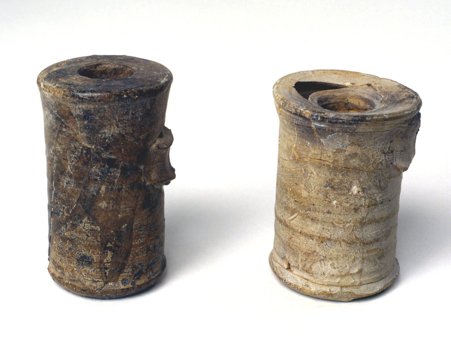Two ancient cylindrical jars with small openings at the top, both weathered and partially damaged, standing upright.