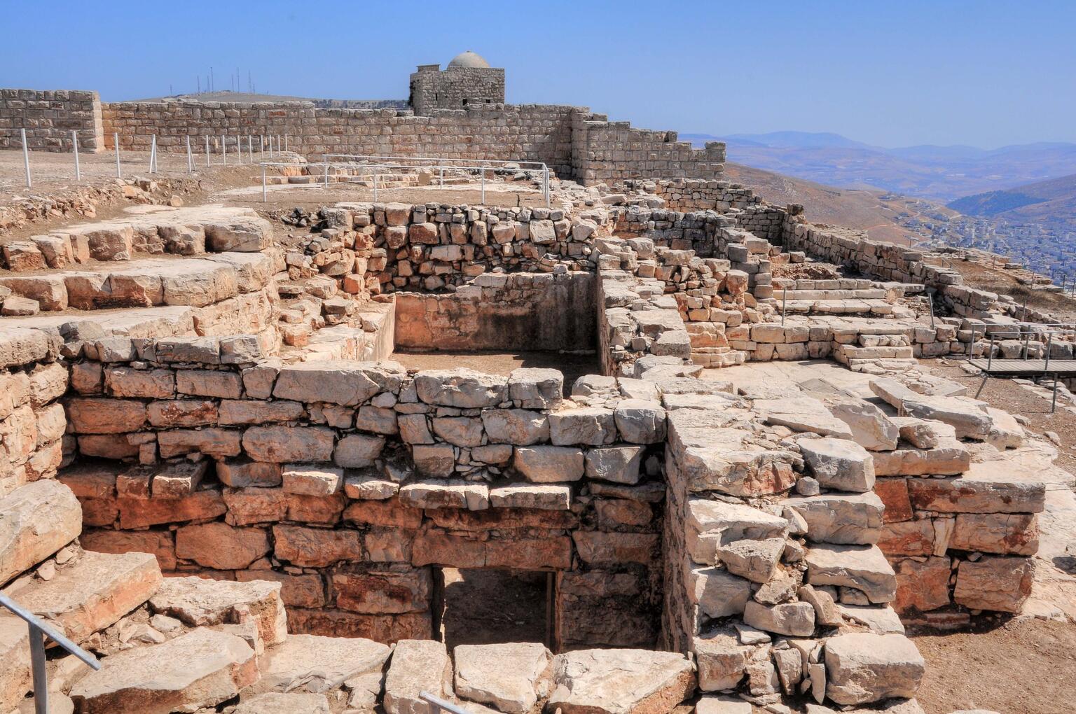 Ancient stone ruins atop a mountain, with multiple walls and chambers visible, overlooking a mountainous landscape under a clear sky.
