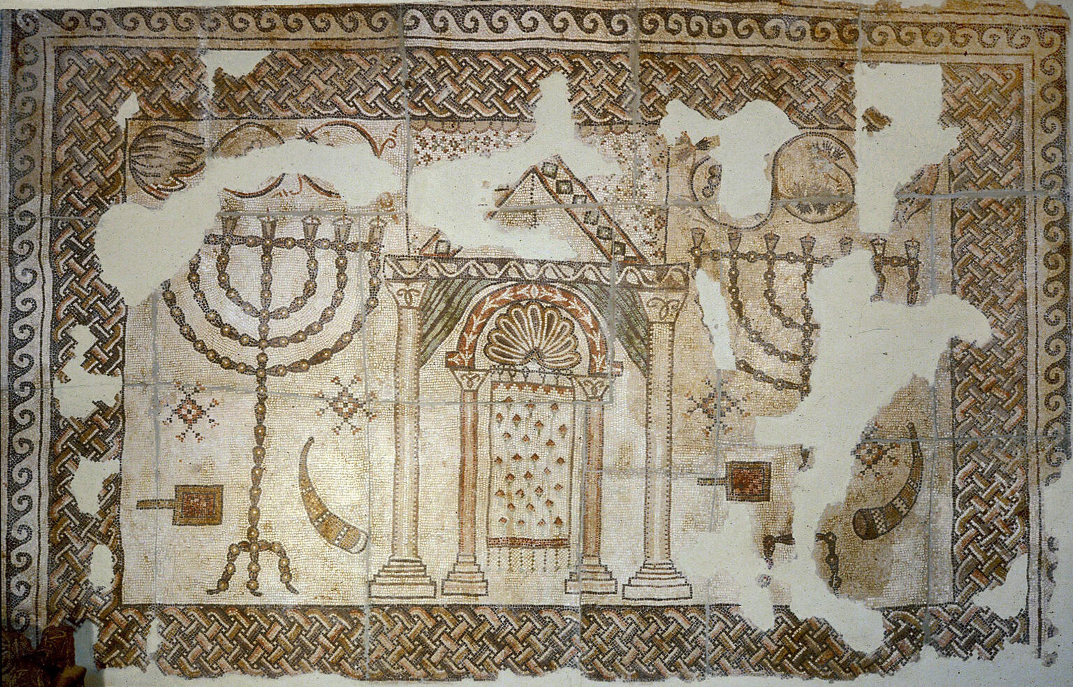 Ancient mosaic featuring a structure with pillars and an arch flanked by two menorahs, each flanked by a shofar and shovel, with intricate geometric borders and some damaged sections.