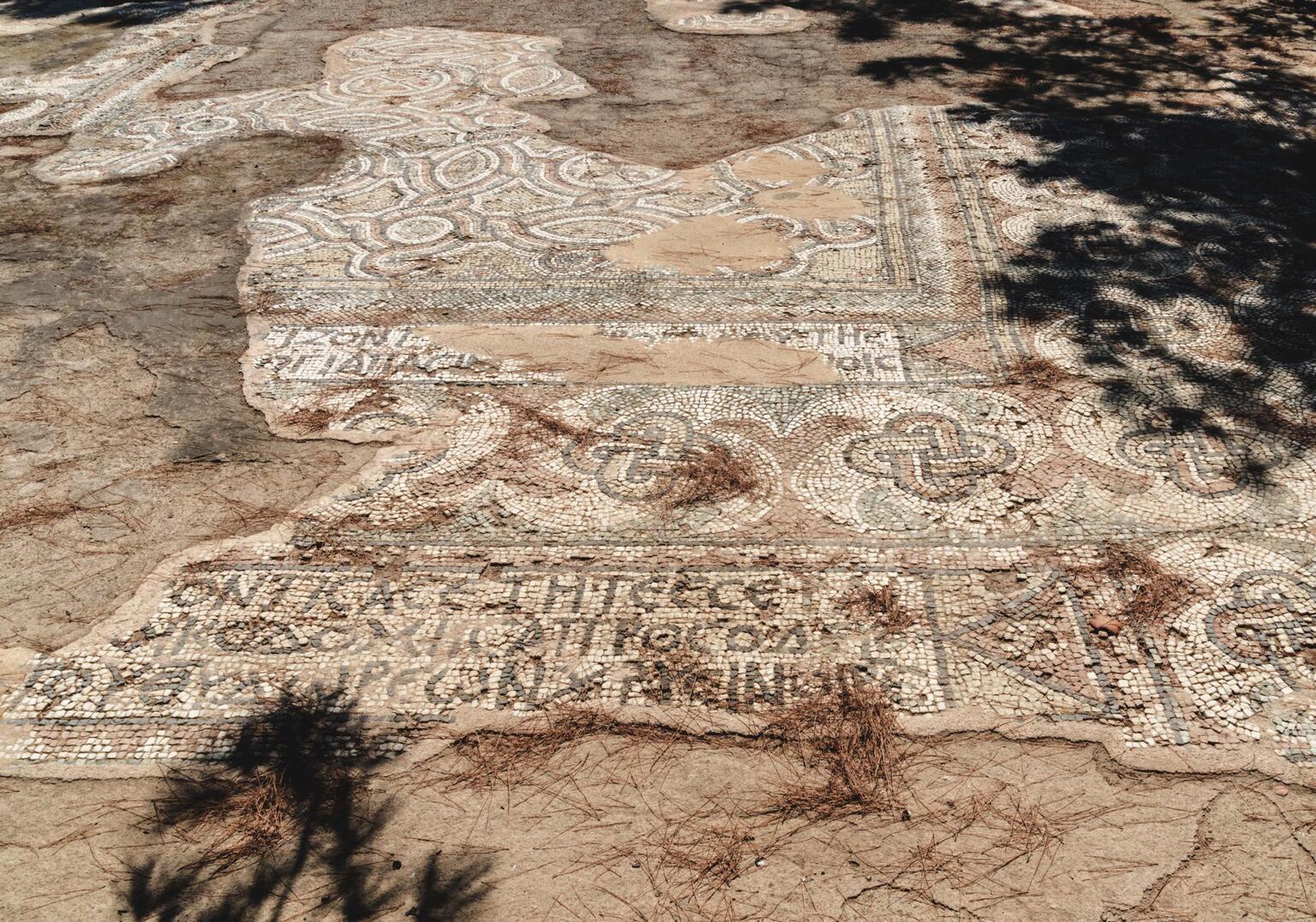 A partially uncovered ancient mosaic on dry, cracked ground, with geometric patterns and some Greek text visible. Shadows from nearby trees fall across the mosaic's surface.