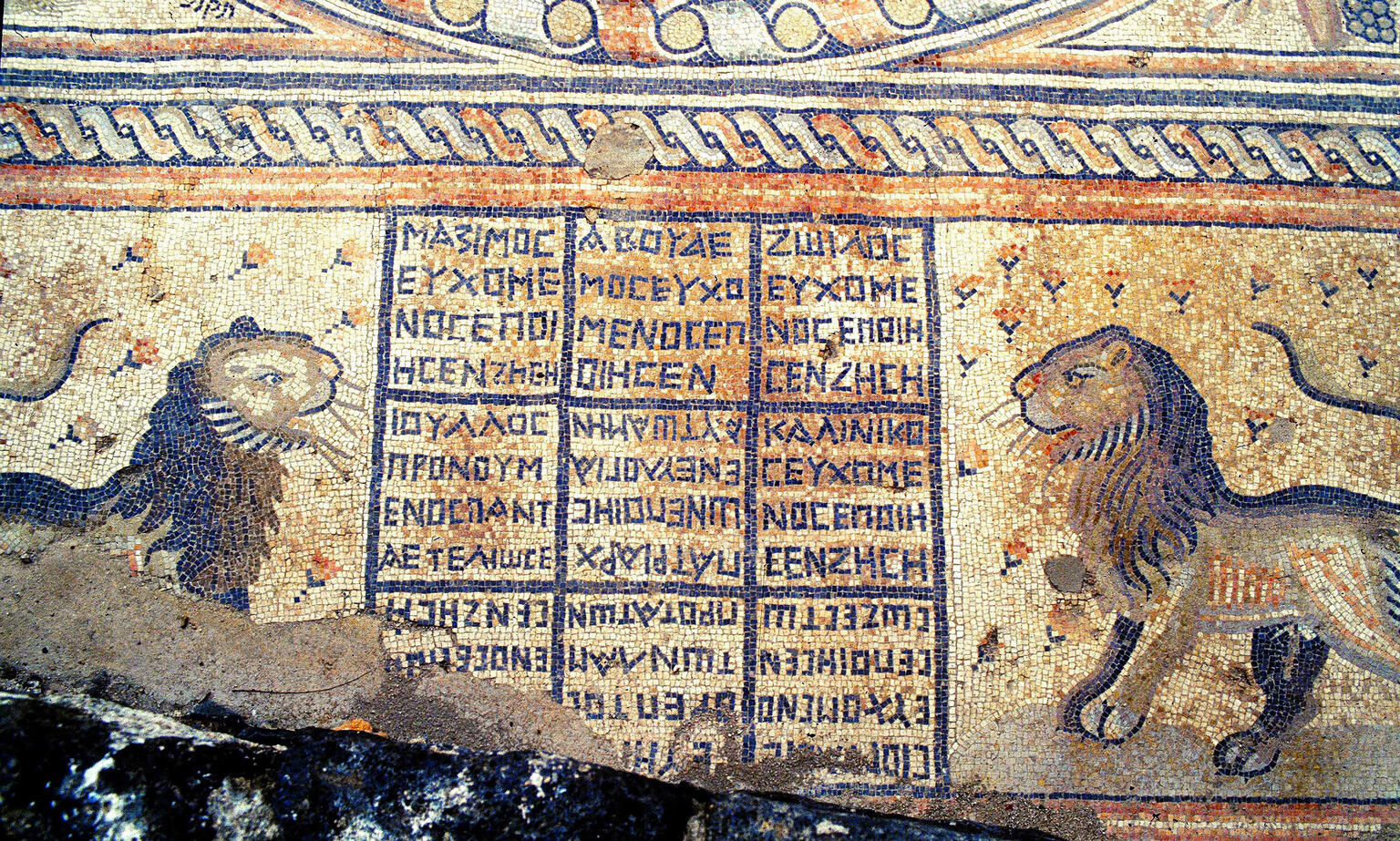 Ancient mosaic featuring two lions facing a central grid with Greek letters, surrounded by decorative patterns. The scene appears weathered, with part of the mosaic damaged in the lower left corner.