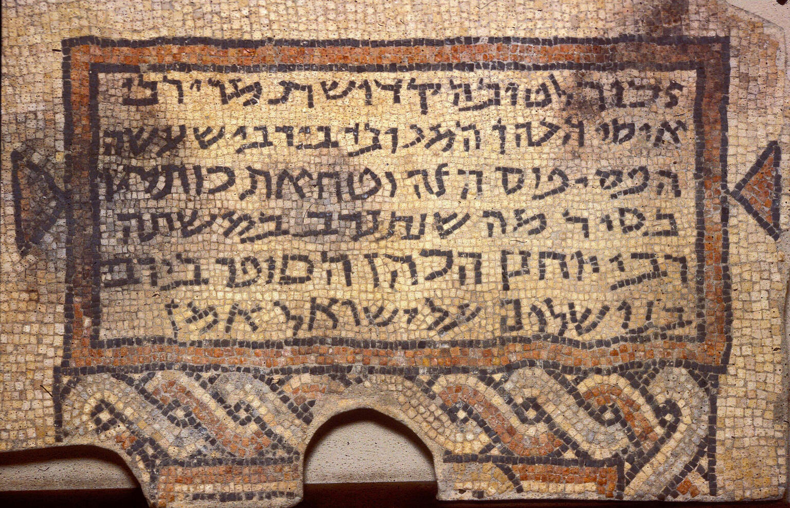 Ancient mosaic with Hebrew script, bordered by geometric patterns, including interwoven designs at the bottom. The text is clearly visible within a rectangular frame.