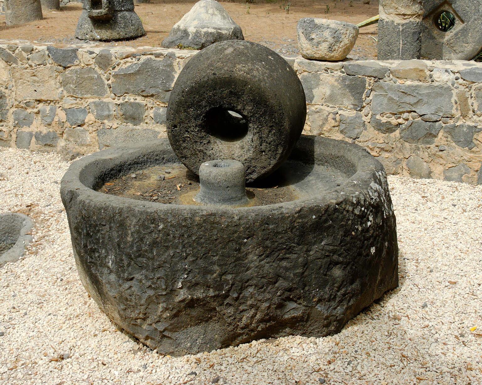 A large, round olive press consisting of a circular basin and wheel sits outdoors on gravel, surrounded by a low stone wall and other stone objects.