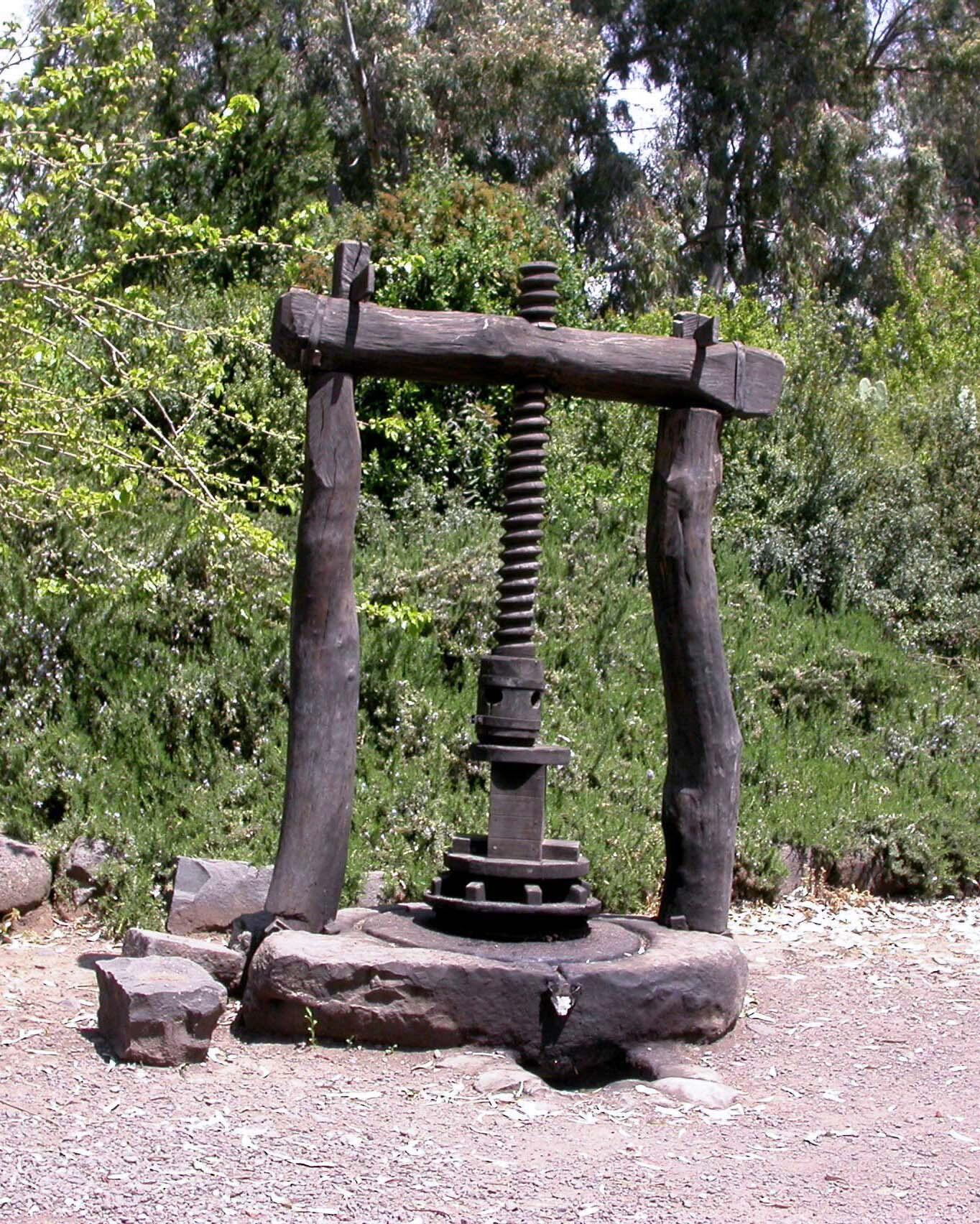 Old wooden olive press with a large vertical screw, set outdoors on a stone base surrounded by trees and greenery.