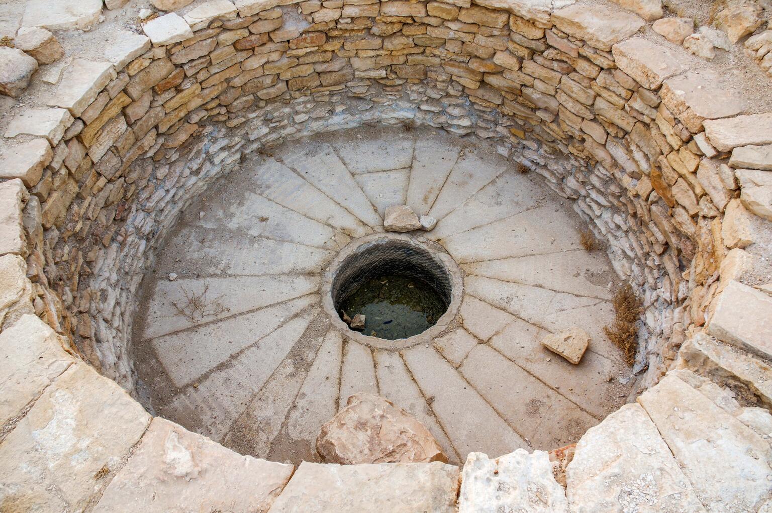 Circular stone basin with layered stone walls, a base with a radial pattern, and a round opening at the bottom. A few rocks are scattered inside.