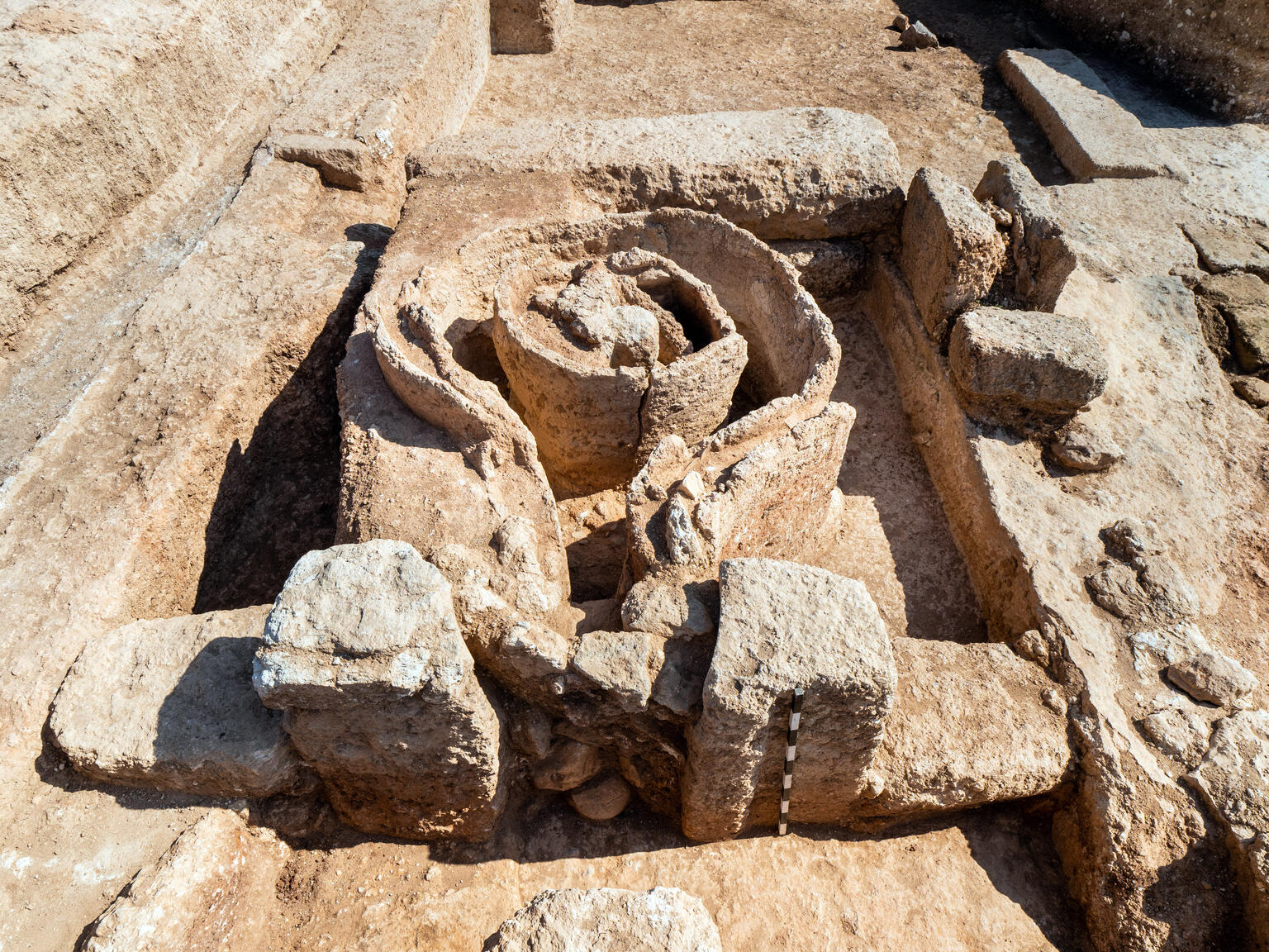 Ancient stone ruins featuring circular and rectangular structures in sandy soil. The weathered stones are arranged in an intricate, partially buried formation.